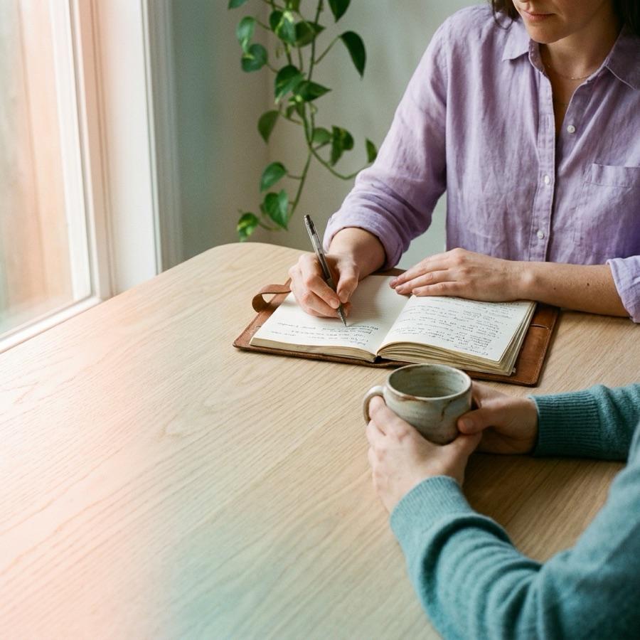 Two people talking across a table in a bright consulting room, one writing in an open notebook, both seen from the chin down