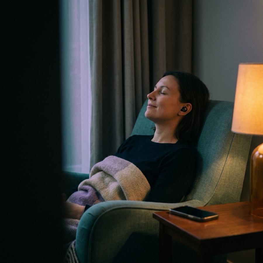 A person reclining with eyes closed in a dim, warm room, listening through a single earphone with a blanket across their lap